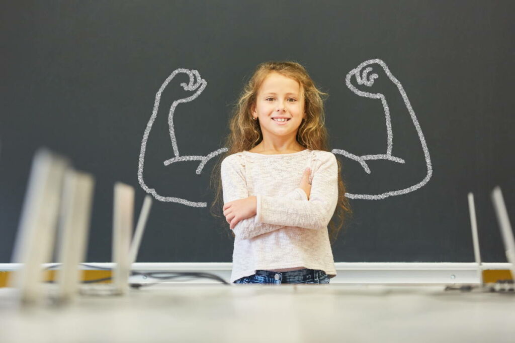 Lerntherapie & Förderung bei Lernschwächen - Lächelndes Mädchen in der Schule steht vor einer Tafel mit Muskeln aus Kreide